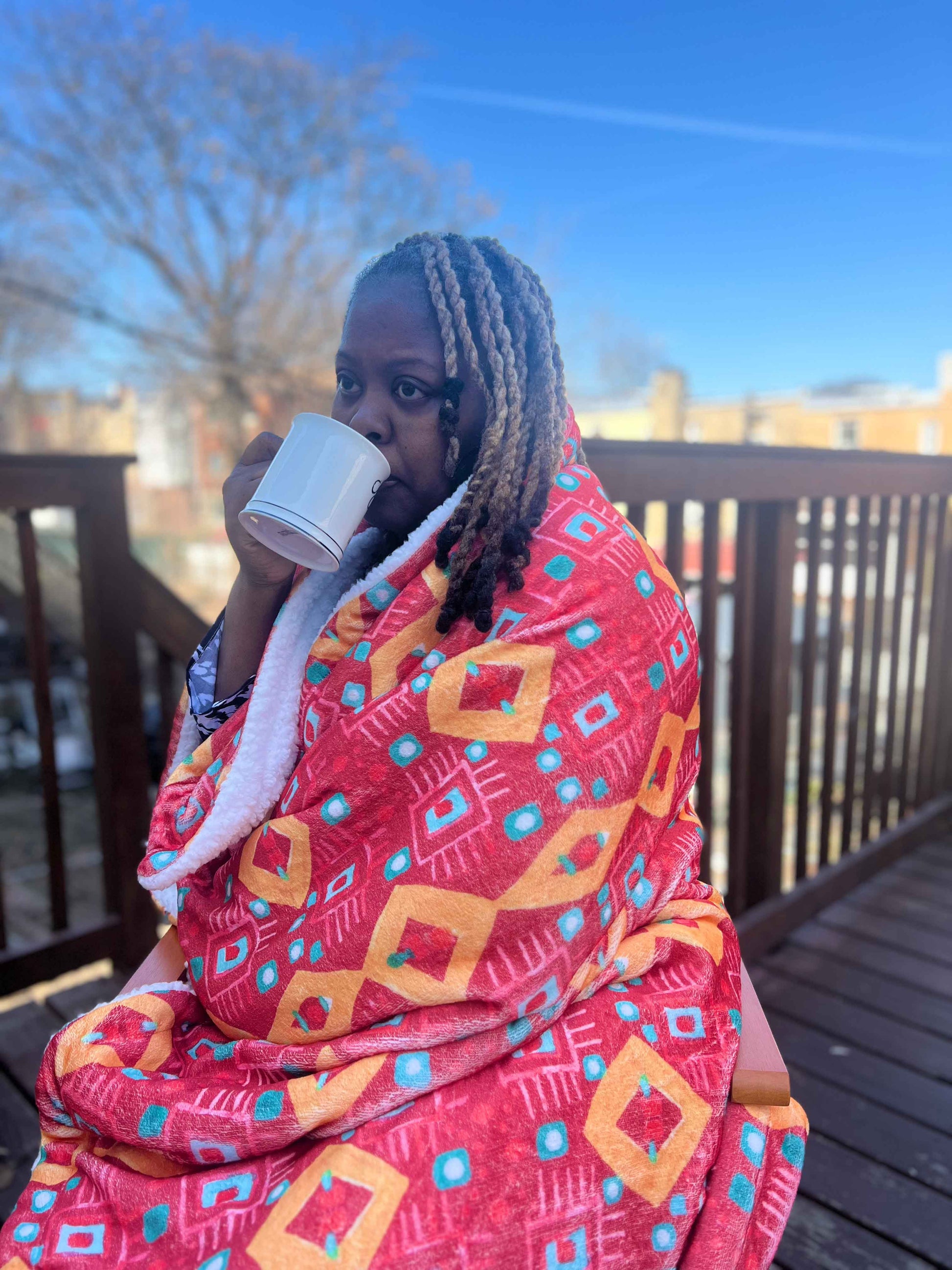 Person wrapped in a red patterned blanket, drinking from a white cup on a deck.