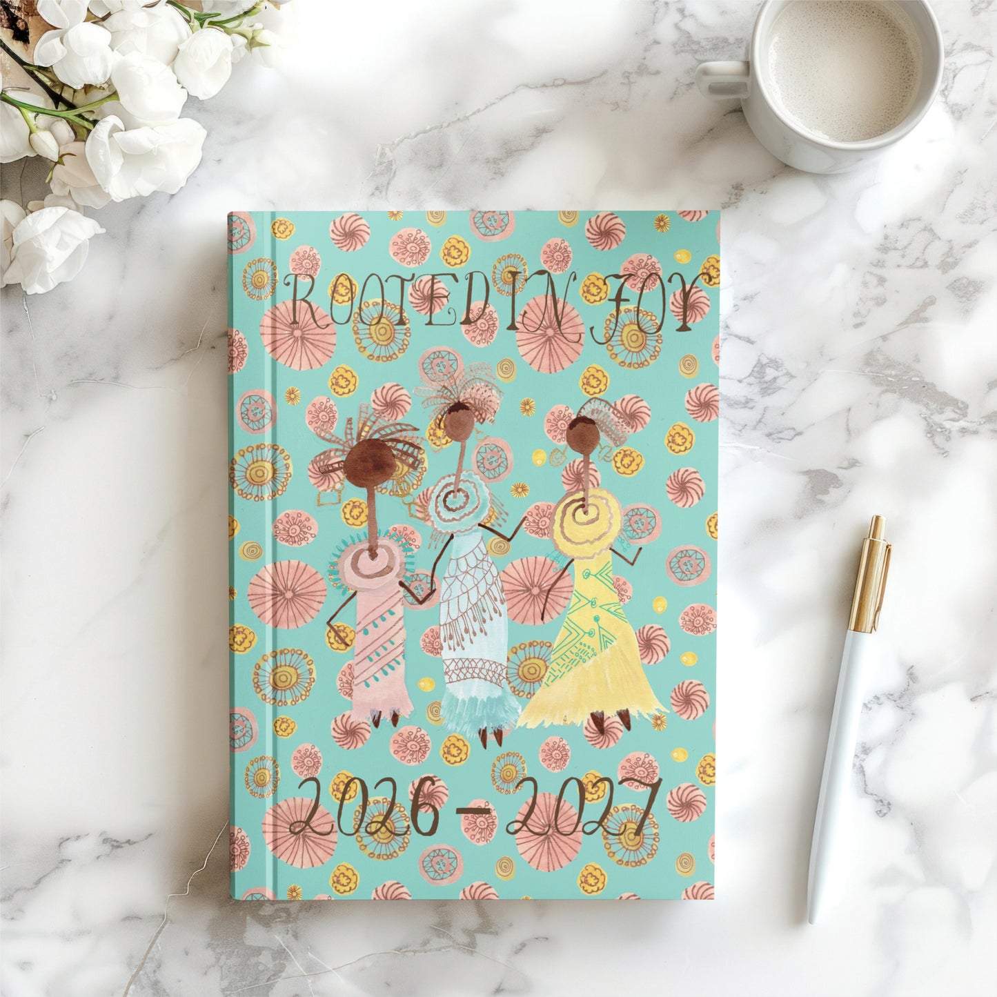 Notebook titled 'Rooted in Joy' on a marble surface with flowers and a mug.