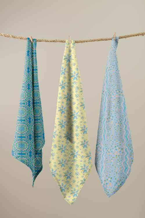 Three patterned kitchen towels hanging from a rope against a plain beige wall. The left towel is blue with a teal circular mandala pattern, the middle towel is pale yellow with a light blue floral snowflake pattern, and the right towel is light blue with a teal wavy pattern.