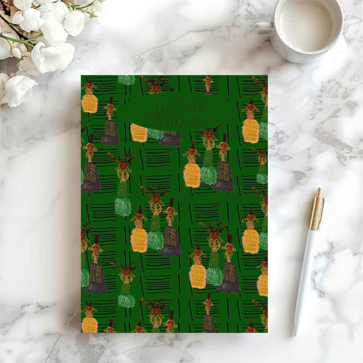 A green patterned notebook or journal cover with illustrated figures and the text 'Bold Thoughts jpurnal' lies on a white marble surface, styled with a cup of coffee, white flowers, and a white pen with a gold cap.