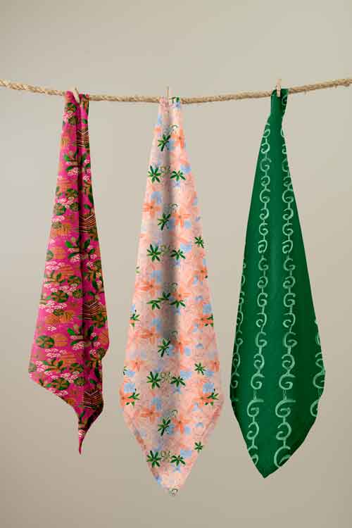 Three patterned fabric cloths or napkins hang from a rope against a plain light beige wall. The left cloth is bright pink with a dark floral print, the middle is pale pink with peach and green palm-like motifs, and the right is dark green with a white chain-link pattern.