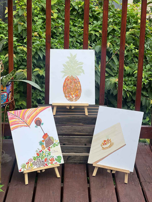 Three original paintings by Lifewithrjarts displayed on small wooden easels outdoors against lush green foliage. The artwork features warm, colorful imagery inspired by nature, food, gathering, and everyday life, including a pineapple and a vibrant market-inspired scene.